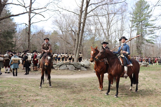 Militia soldiers mounted on horses look off into the distance. Lines of militia soldiers stand in the background.