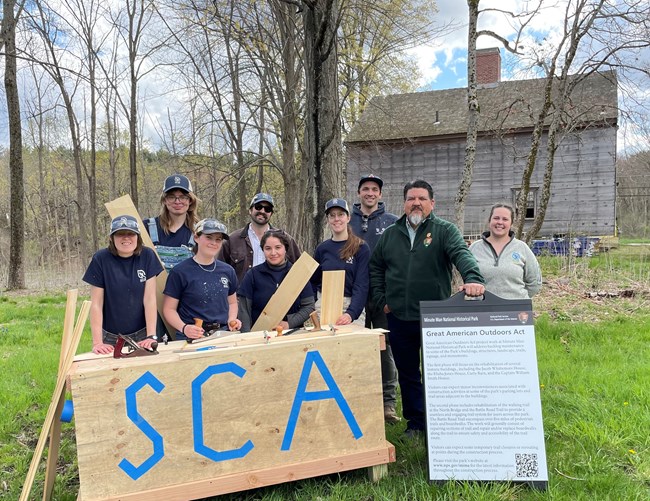 Director Sams standing with SCA members behind a work bench in front of a historic house