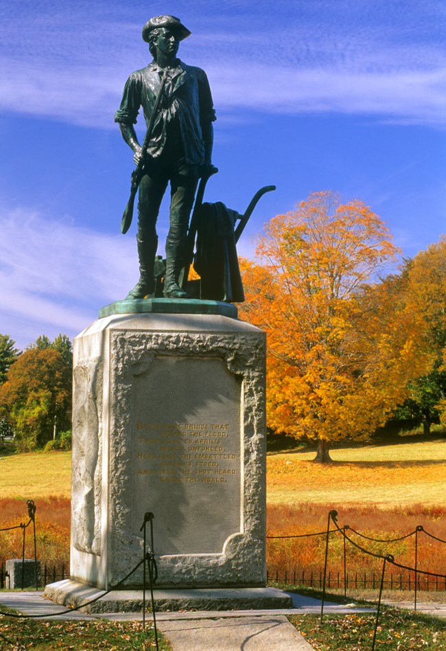 Bronze statue on a granite base depicting a Revolutionary War soldier with musket and plough. Autumn leaves in the background