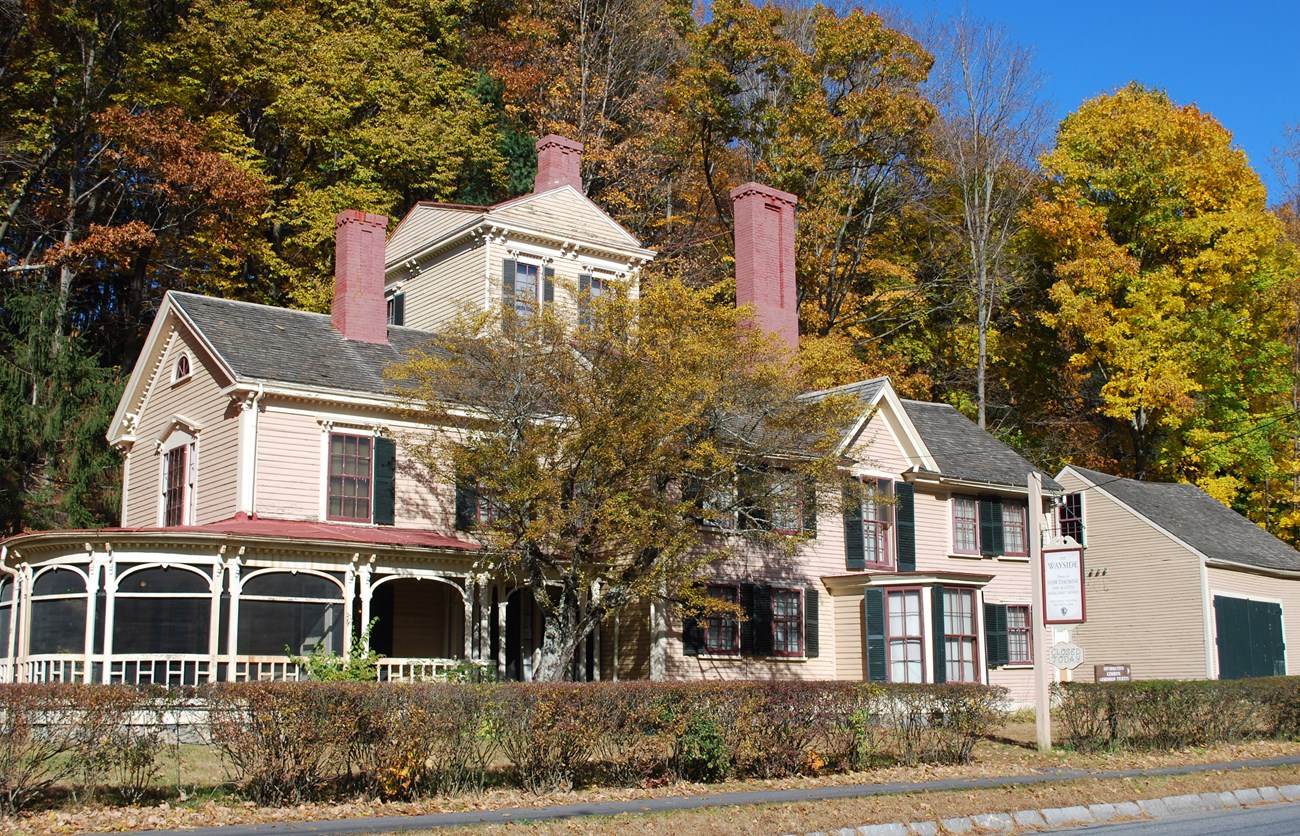 A large wooden house painted yellow. The home is 6 bays wide. The main structure is two stories with a three story tower in the center. A large porch is attached on the left side. In the background a hillside is covered in trees.