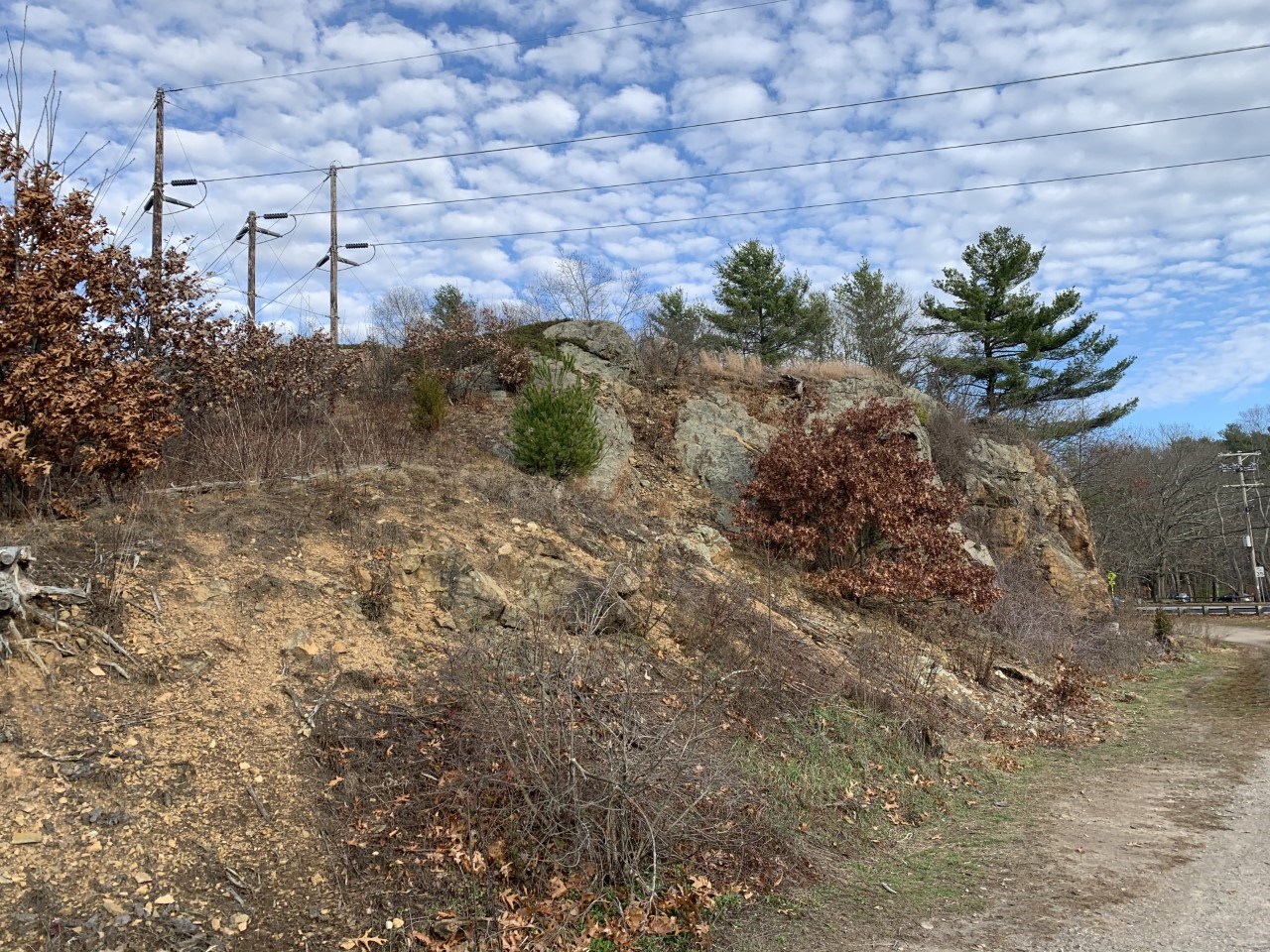 A sharp outcrop of red/tan rocks ascends skyward with small bushes scattered around its base. A stone monument stands at the center bottom of the cliff face. Trees tower on either side of the hill