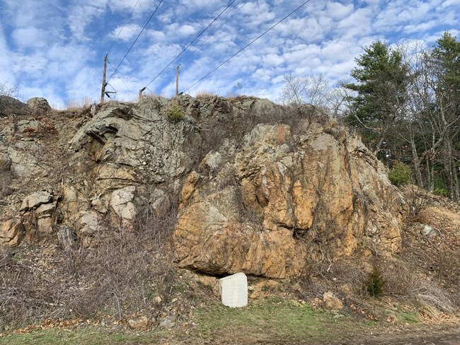 A sharp outcrop of red/tan rocks ascends skyward with small bushes scattered around its base. A stone monument stands at the center bottom of the cliff face. Trees tower on either side of the hill