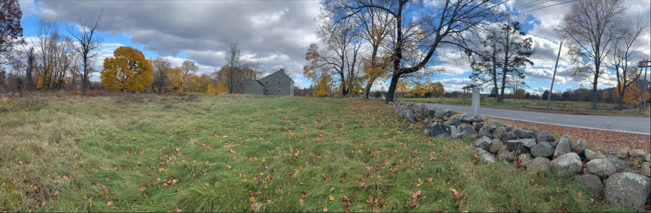 A road with stone wall runs along the right side of the image. The road is through a meadow surrounded by trees in the distance. A wooden historical house is at the center front of the scene.