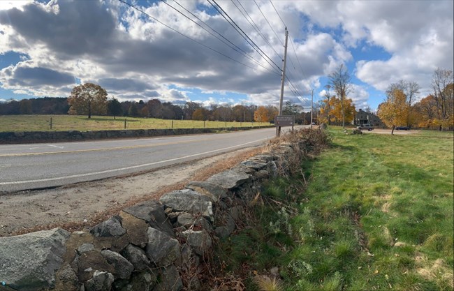 A stone wall lines a modern road. The road cuts through an open meadow with green grass on either side. In the distance trees cover a hilltop. A parking lot on the right of the road is marked with a historical sign.