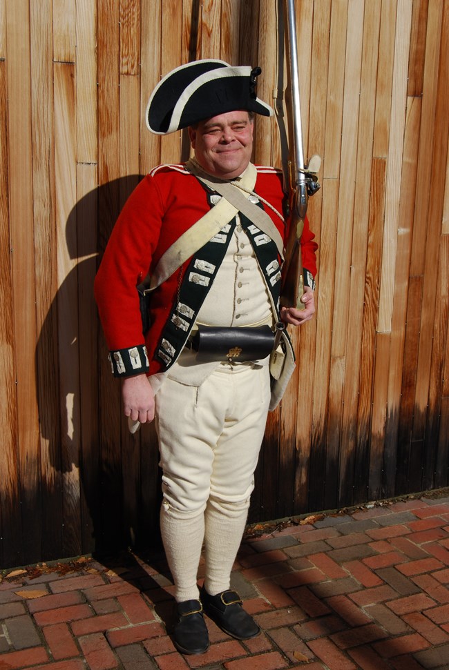 Man dressed as Revolutionary War British soldier with red coat, cocked hat, armed with a musket and bayonet standing in front of a wooden building.