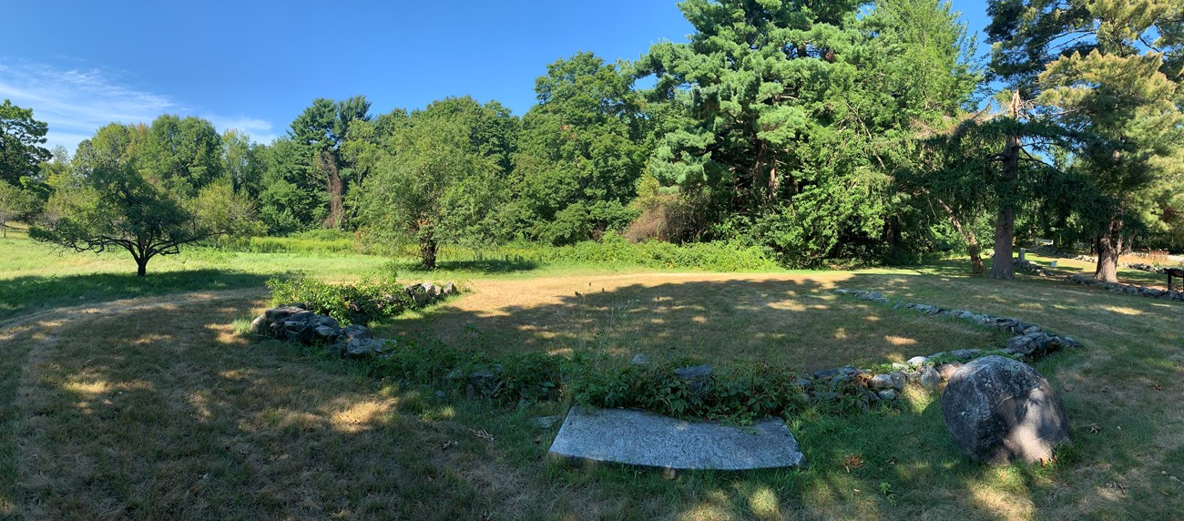 A low stone foundation marks three sides of a historic building site in an open grassy field. In the distance a hill is obscured by a thick layer of green leafy trees.