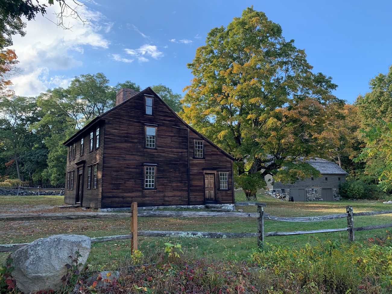 A side view of a two story salt-box style house made of brown wood. A large tree towers over the rear of the house and a wooden barn in seen in the distance. orange, red, and brown leaves indicate autumn.