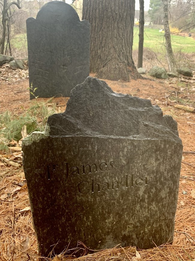 A small slate foot stone stands in the foreground of the main headstone. This footstone marks the end of the grave and reads “Mr. James Chandler.”