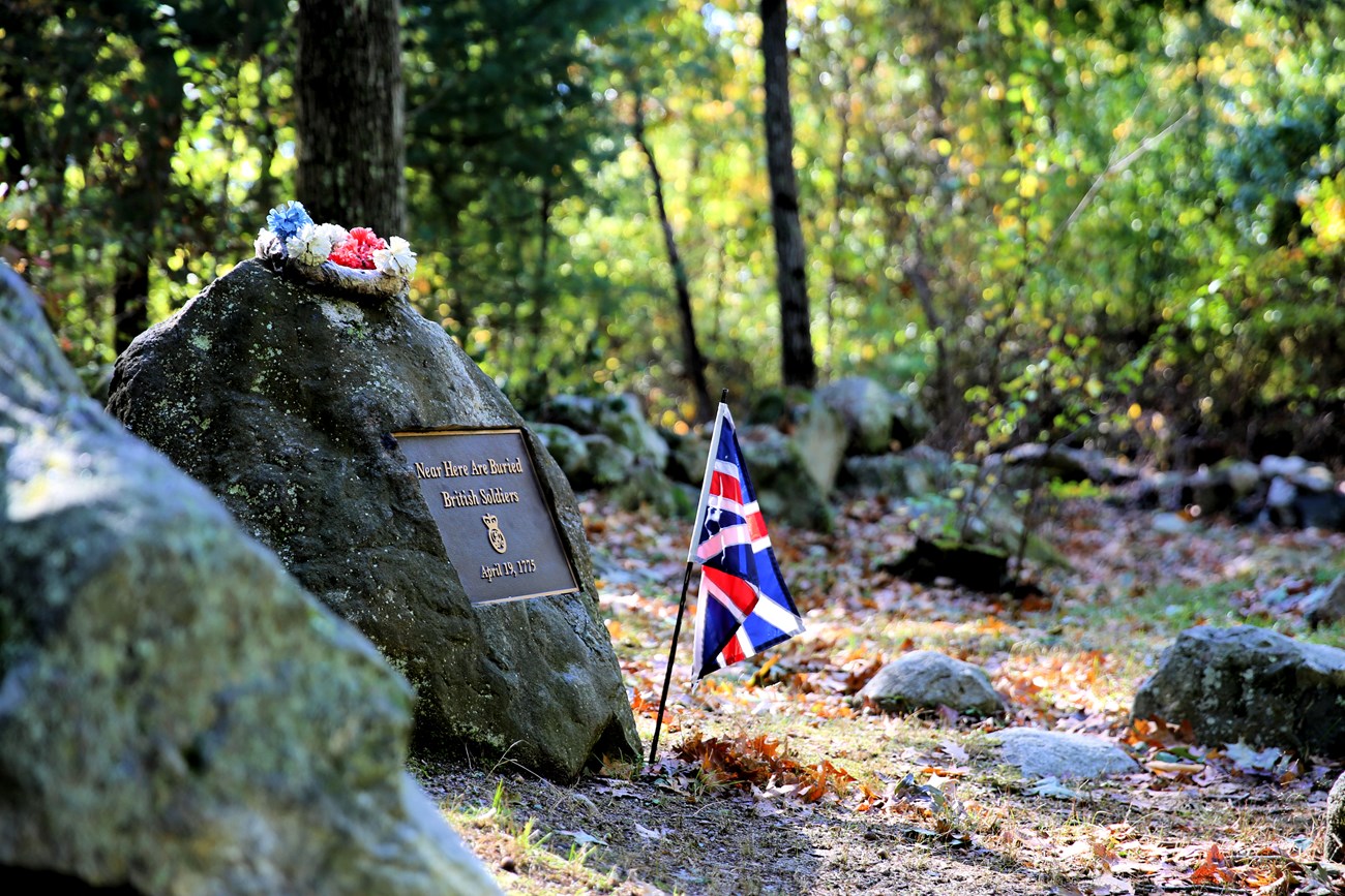 A grave stone with metal plaque inset sits off to the left side of a dirt road. Behind the stone is a thick tree line and a stone wall. A small wreath of red, white, and blue flowers sits atop the stone and a small British flag is planted at its base.