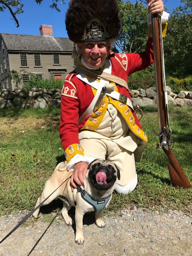 Man dressed as Revolutionary War British soldier with a red coat, black fur cap and a musket, kneeling down and posing with a small dog