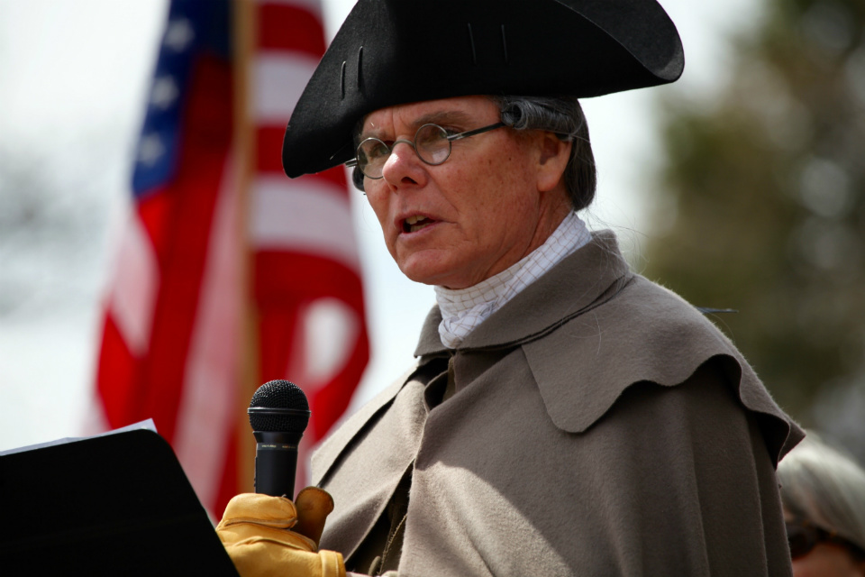 Volunteer Ed Hurley speaks to people about sacrifice. He is dressed in colonial clothes, wearing a black tricorn hat and a buff great coat. Behind is the American flag. Speaking of sacrifice, volunteer Ed Hurley, dressed in 18th century clothes speaks of the sacrifice of veterans.