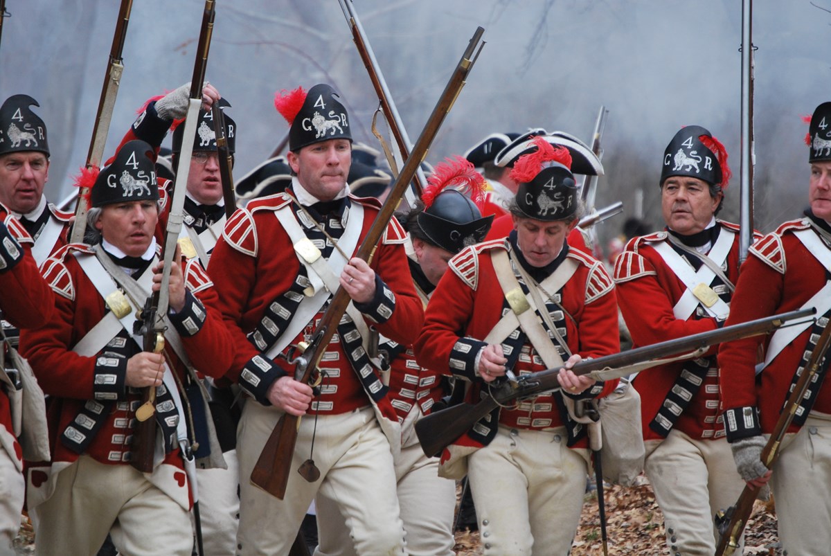 A group of 8 British Revolutionary War soldiers in red coats and black caps with muskets march together through a cloud of gunpowder smoke