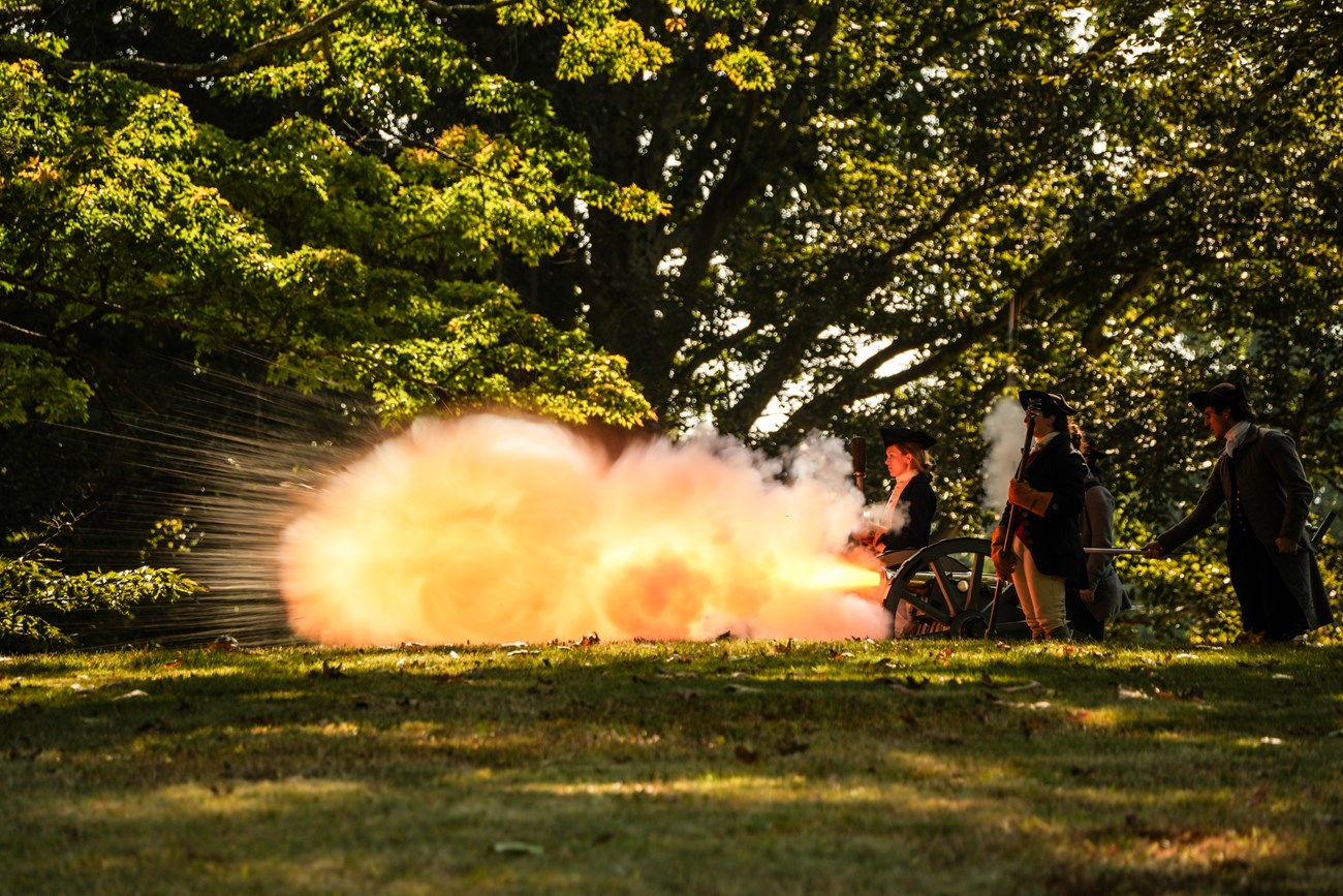 A large flame shoots from the muzzle of a cannon as the fire illuminates the faces of the crew nearby