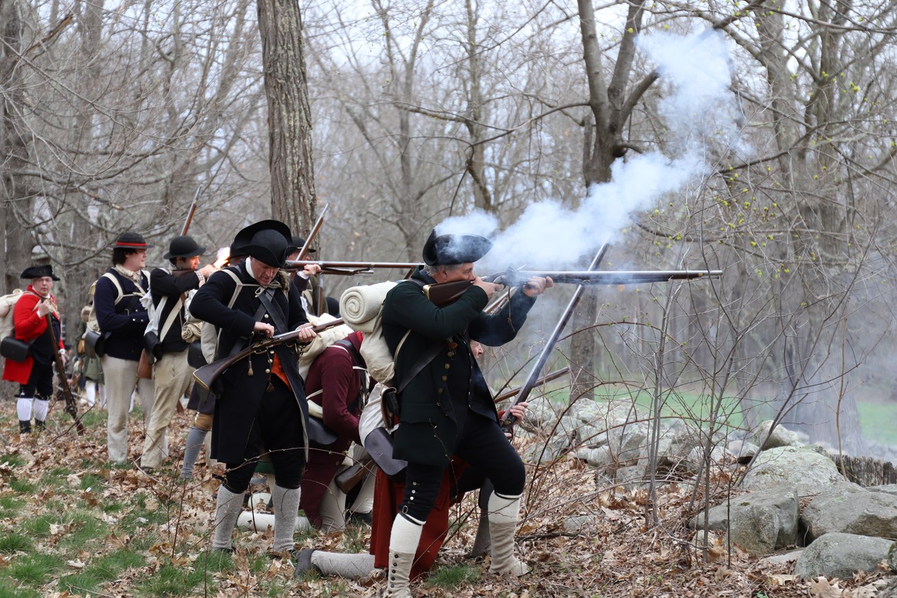 Colonial militiamen fire their muskets from behind a stone wall.