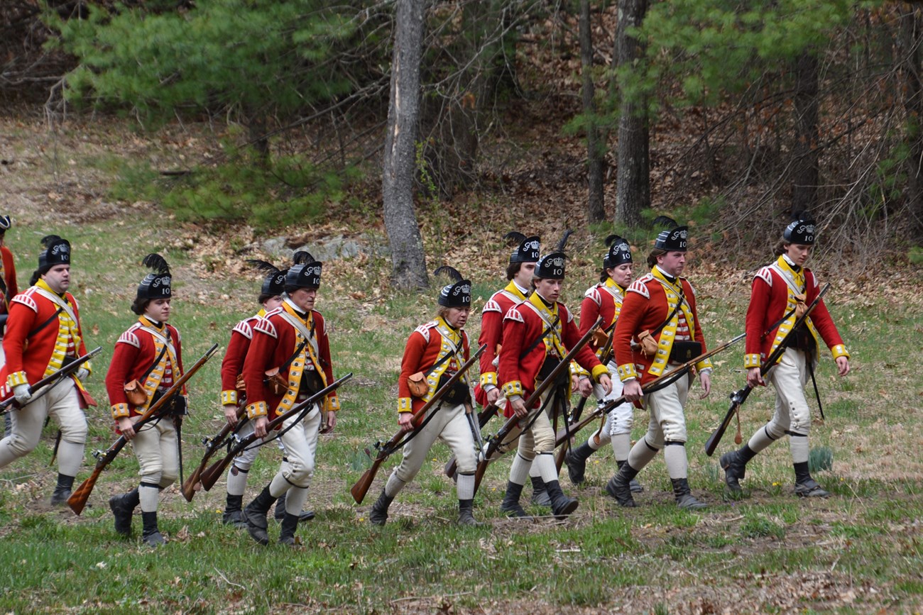 British soldiers in 1775 light infantry uniforms march in open order through a field surrounded by trees