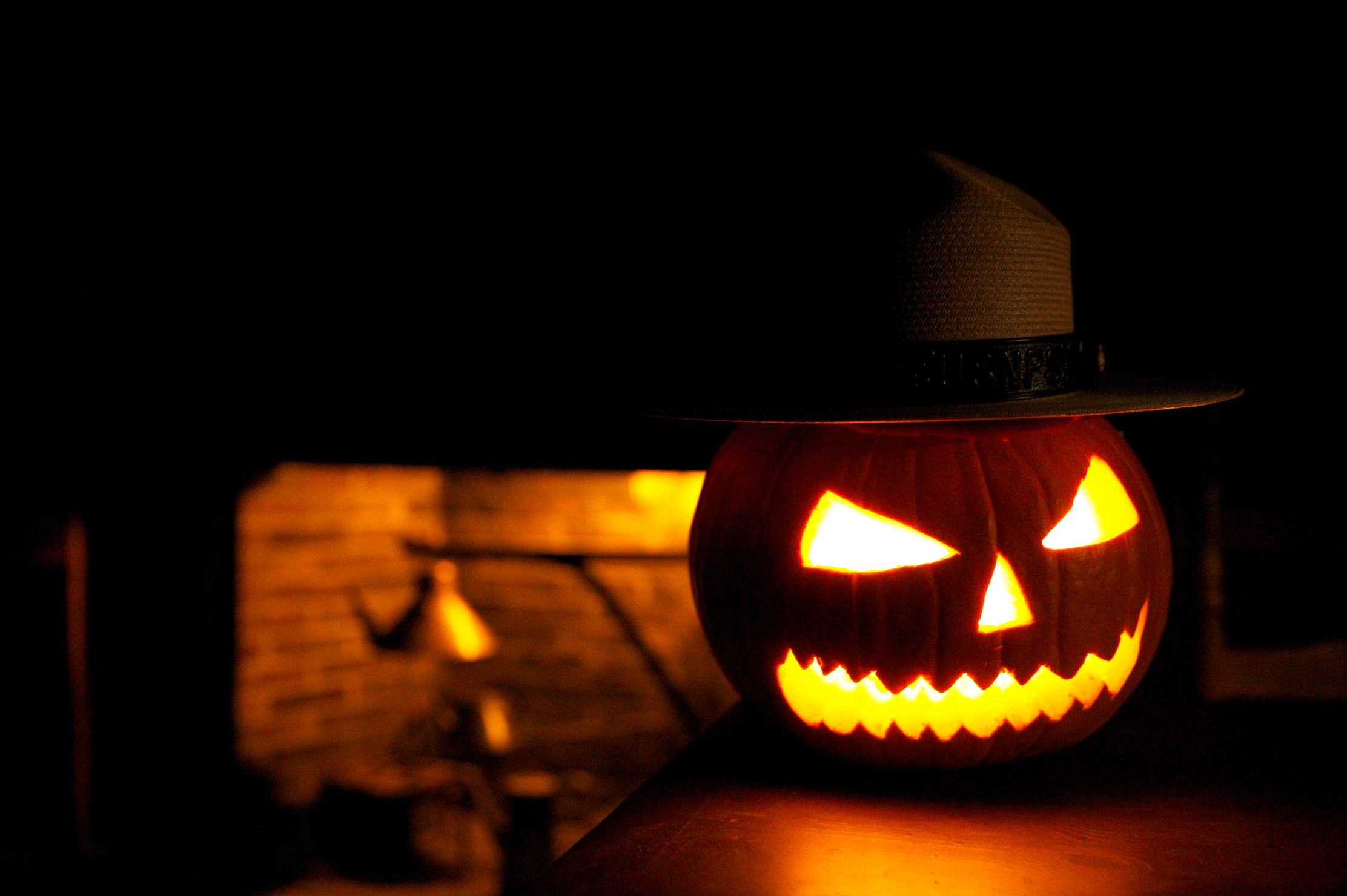 Flat Hat Pumpkin A pumpkin carved with a grinning face illuminated by an interior light. An NPS Flat hat sits on top. In the background a fireplace is illuminated in a dark room.