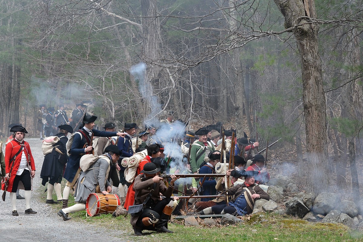 A group of militia soldiers take cover behind a stone wall. Some are firing muskets