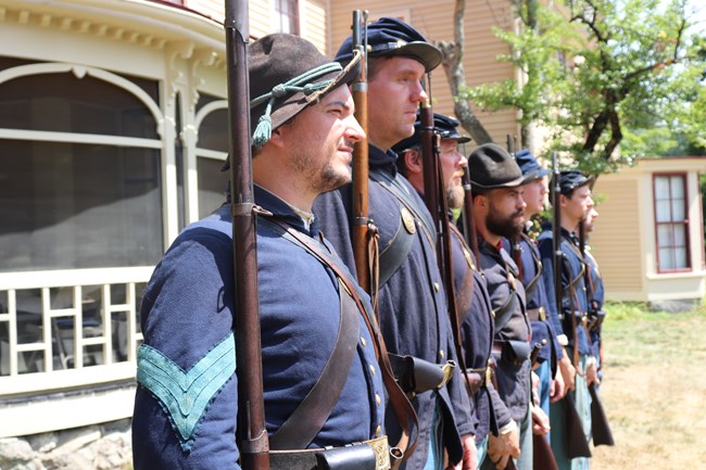 A group of living history volunteers in Union Army Uniforms stand in a row with the yellow siding of the Wayside House behind.