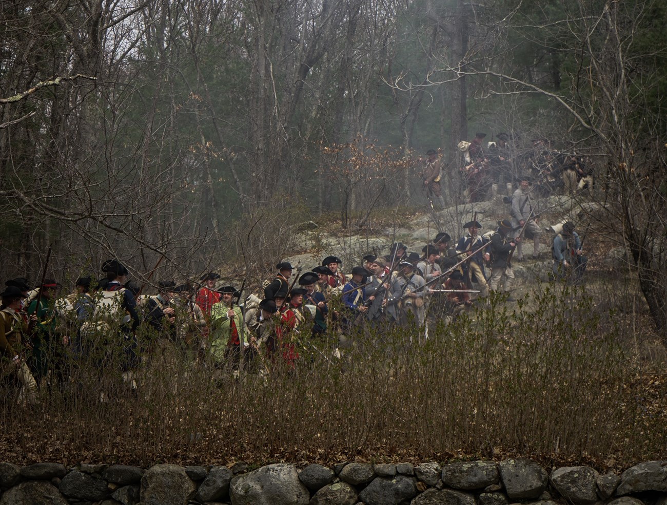 Colonial militiamen stand in line in a wooded landscape and fire their muskets