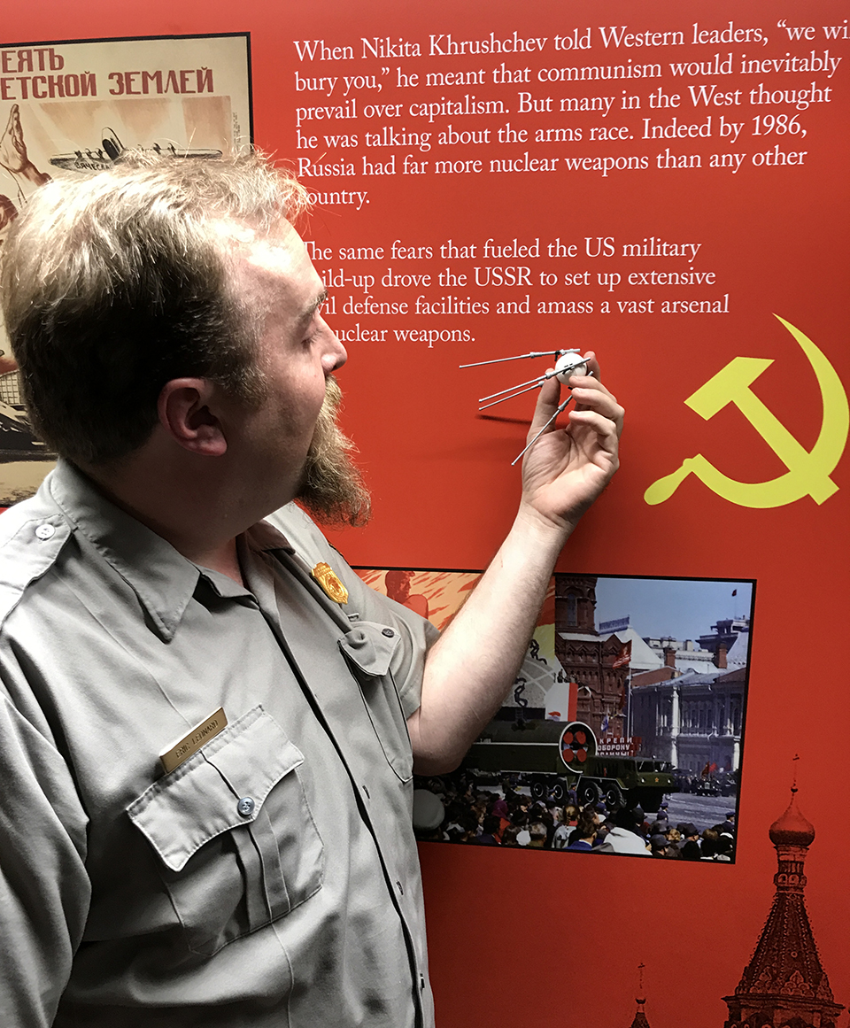 Superintendent Eric Leonard displays a LEGO Sputnik model in front of an exhibit panel. Park ranger holds a LEGO model of Sputnik in front of a red exhibit panel