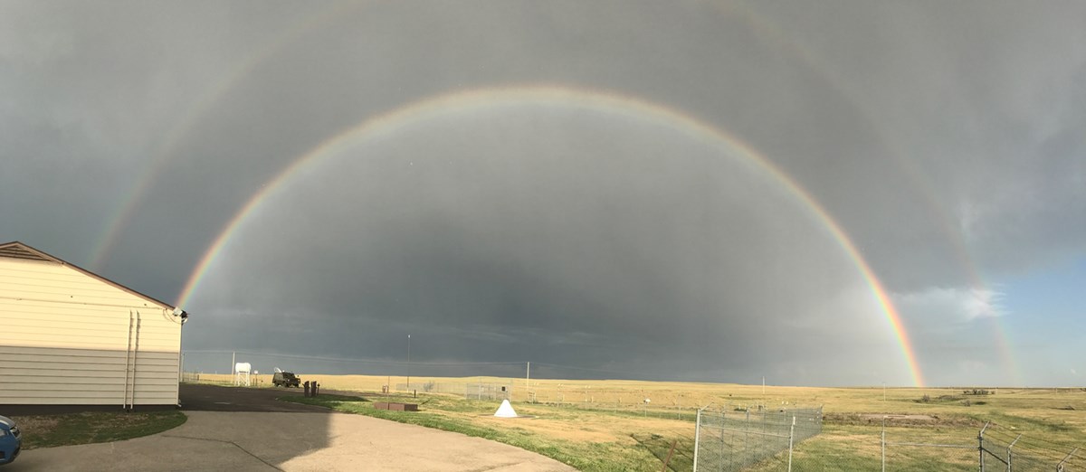 A double rainbow stretches over a prairie landscape with a fence and building in the foreground