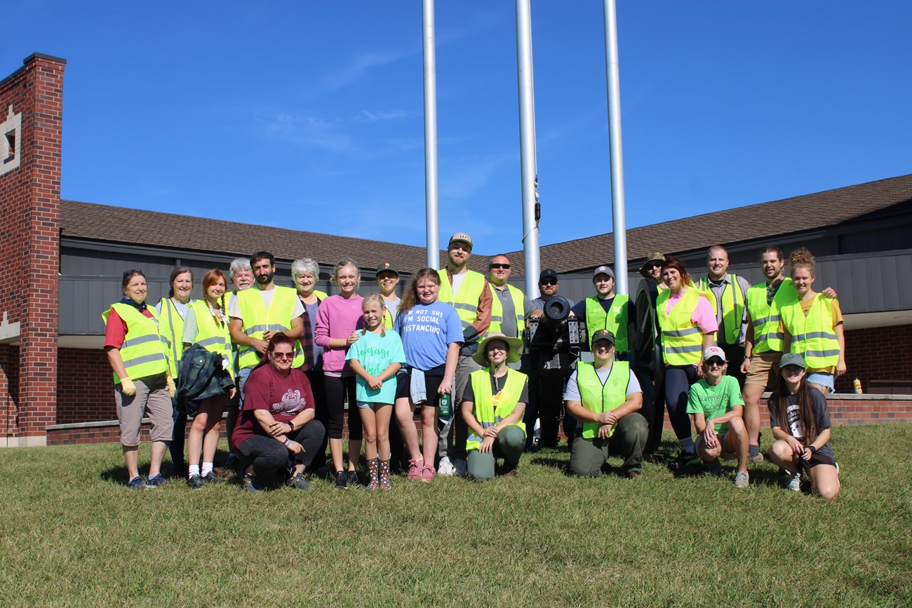 A group of people wearing yellow reflective vests posing for a picture around a cannon in front of a brick building.