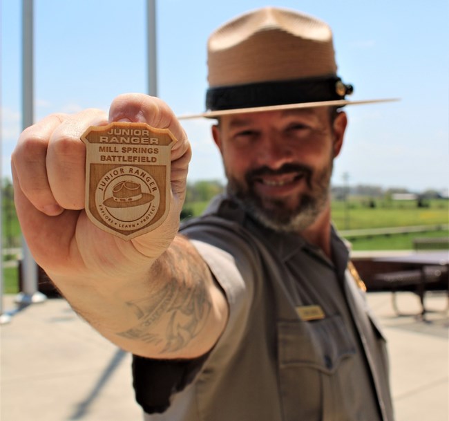 Park Ranger in full uniform holding Junior Ranger Badge up to the camera.