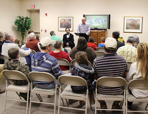 Man at podium speaks to room full of people seated in folding chairs