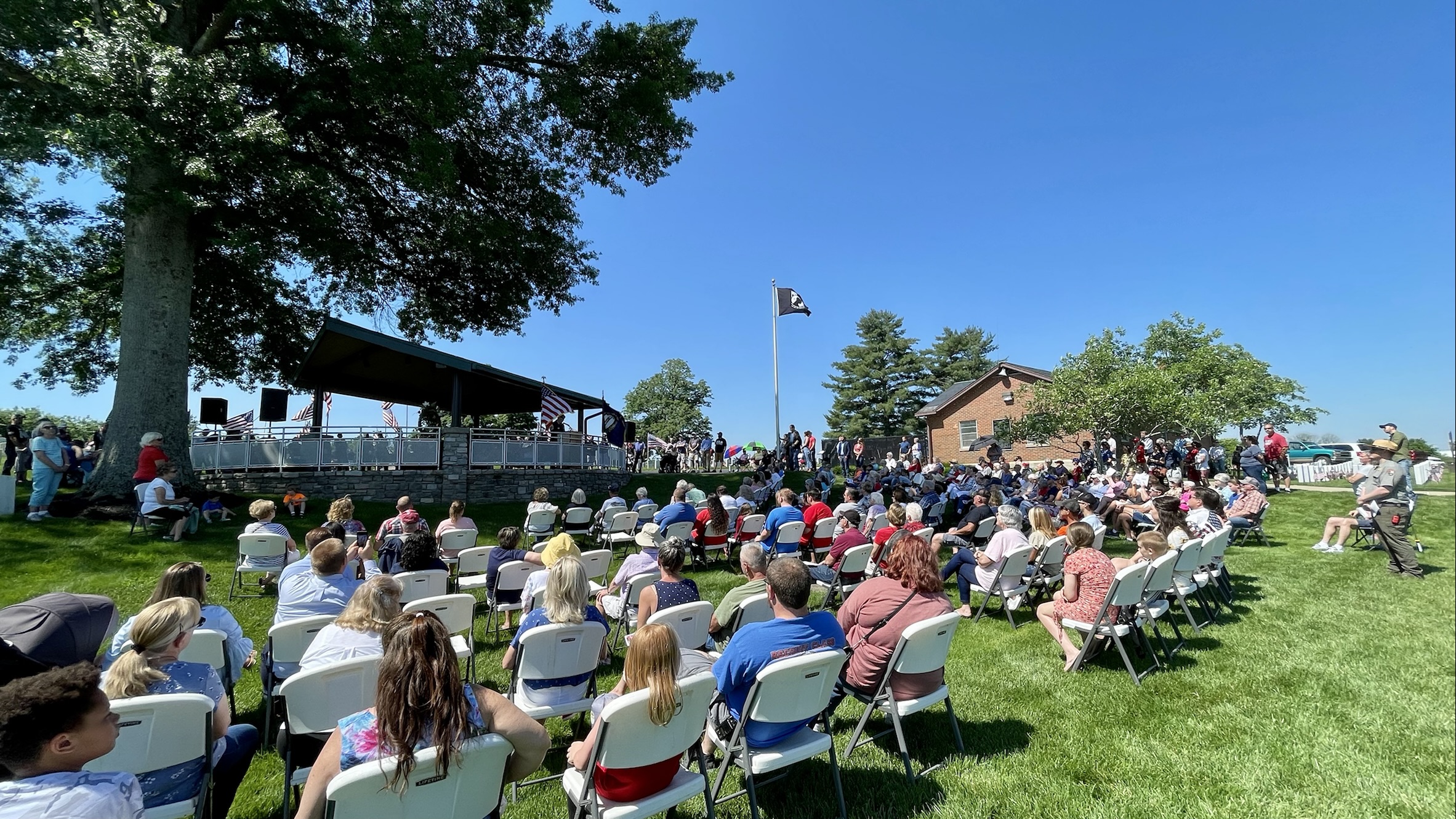 A crowd of people looking at a covered gazebo with a speaker at the podium. Multiple US flags, a Kentucky flag, and a POW-MIA flag are flying against the backdrop of a clear blue sky.