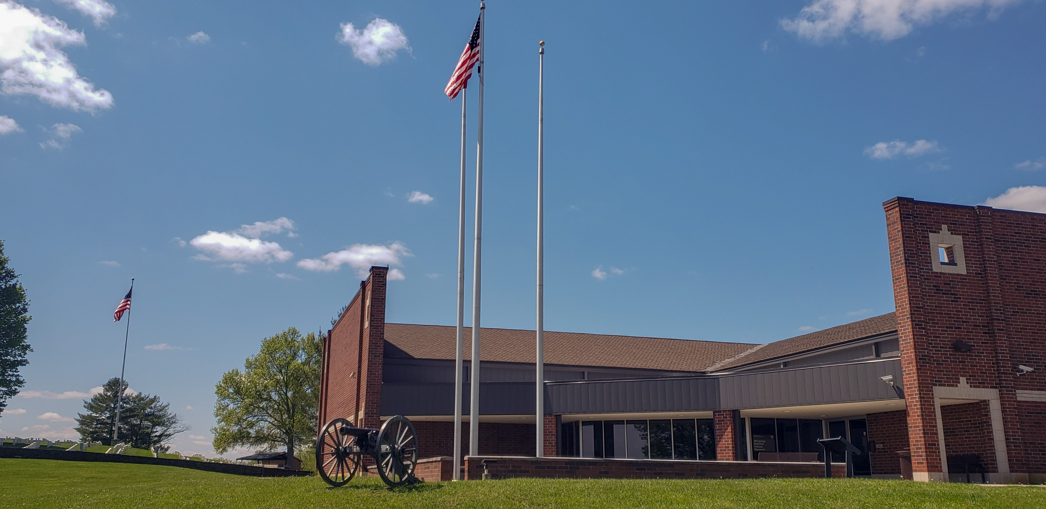 A square red brick building with three flag poles in front, one with the American flag waving and a black cannon underneath.