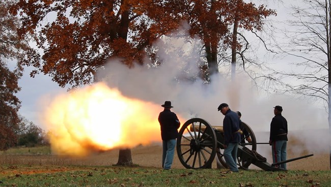 Four people in Federal uniforms stand in proper safety positions around a Civil War cannon as it goes off.