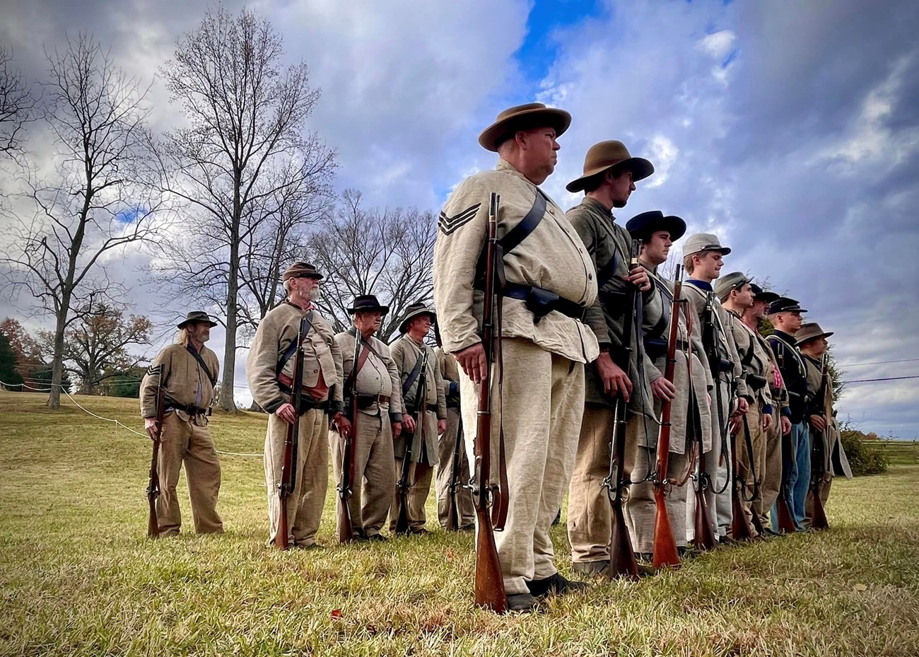 A group of infantrymen wearing grey Confederate uniform lined up, holding their muskets.