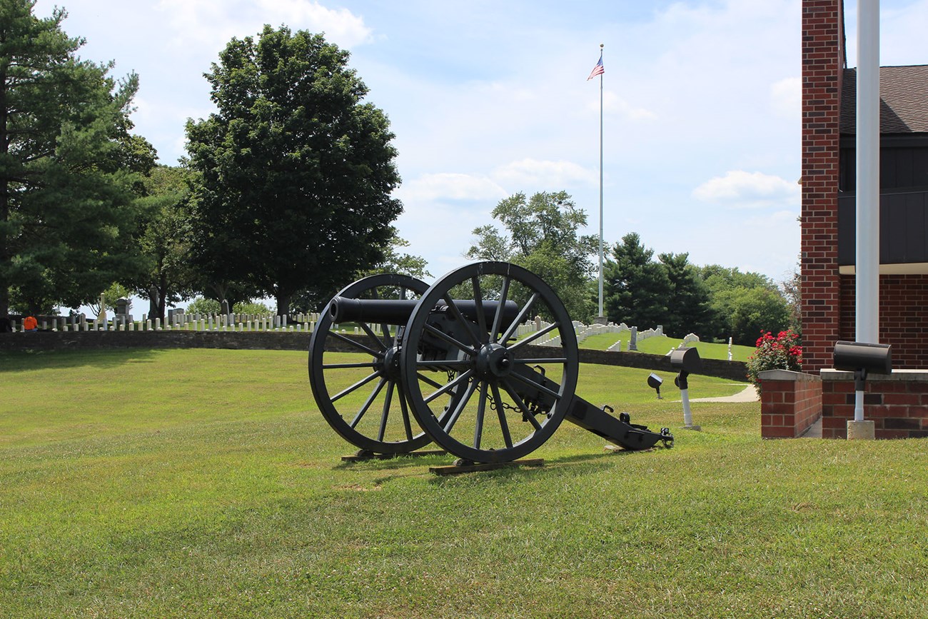 A cannon rests on the grass next to a brick building with national cemetery in background