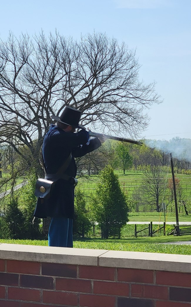 A reenactor wearing blue Civil War uniform and black hat fires musket