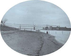 A man sits on the bank of a large river. A steel structure bridge crosses the river in the background.