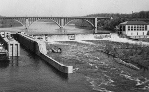 A lock and dam span the river. A large concrete bridges crosses the river in the background.