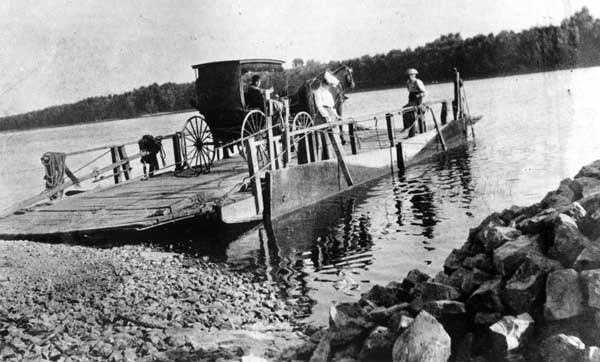 A carriage and horses are loaded onto a rickety wooden ferry to cross a large river.