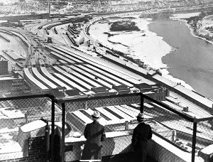 Two men look down on a snowy rail yard full of trains from a bluff top.