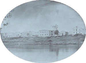 Buildings sit on the top of a low bank. A river runs in front of the town.