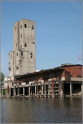 A blue sky and the Mississippi River frame the elevator and sackhouse while piers rise from the water around the structure.