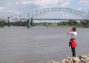 A person uses binoculars to scan the river for birds. A large bridge spans the wooded shorelines in the background.
