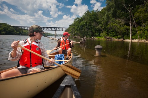 Canoeists boat past a shallowly-submerged cement wall and a metal fixture.