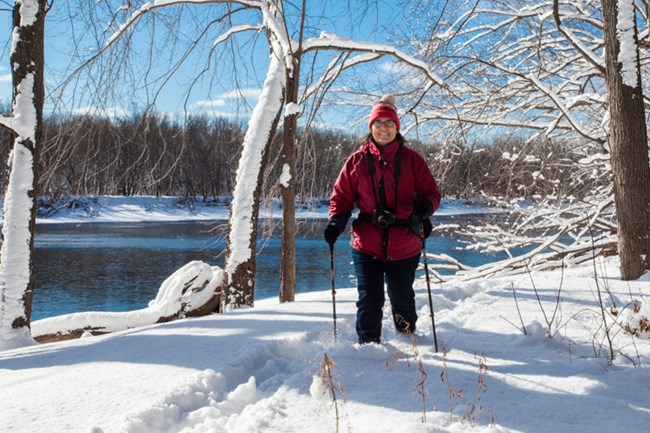 A lone figure in a red coat hikes in a snowy forest beside a river.