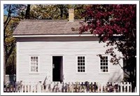 An old small white house with grey roof and a tree with red leafs in foreground