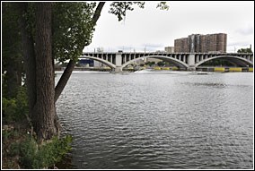 The Mississippi River flows past a wooded island shoreline and towards a city skyline.