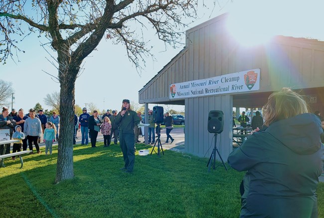 A park ranger speaks into a microphone in front of a crowd, behind her are speakers, a picnic shelter, and banner.