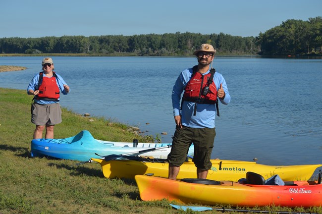 Two people wearing life jackets and smiling stand beside kayaks that are lined up along the shore.