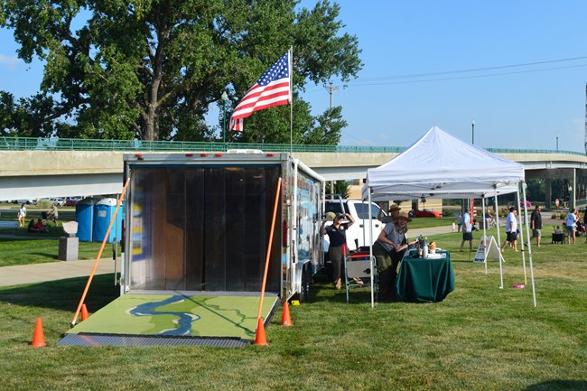 The back entrance to a trailer is open, beside it are tables and pop-up tents set up at an event.