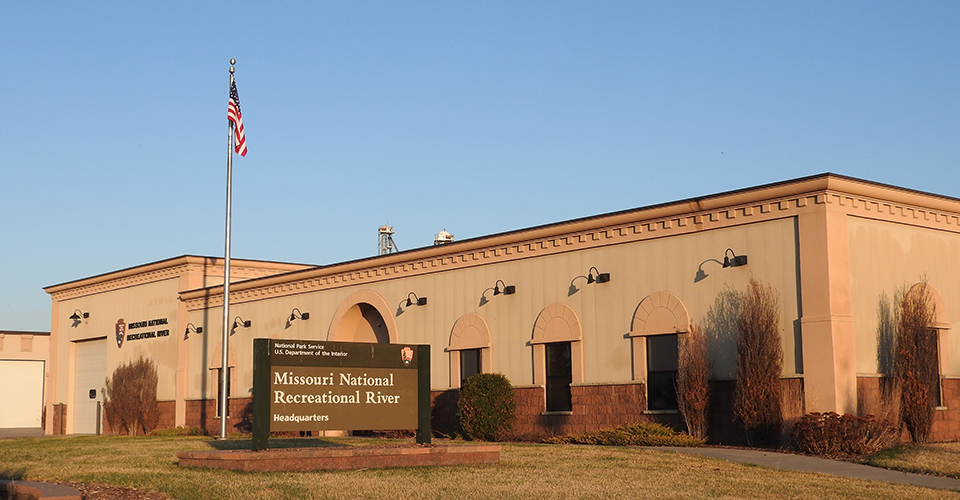 mnrrhq A large sign stands before a tan, one-story building. The sign reads Missouri National Recreational River. A silver flag pole located at the front entrance to the building with an American flag is behind the sign.