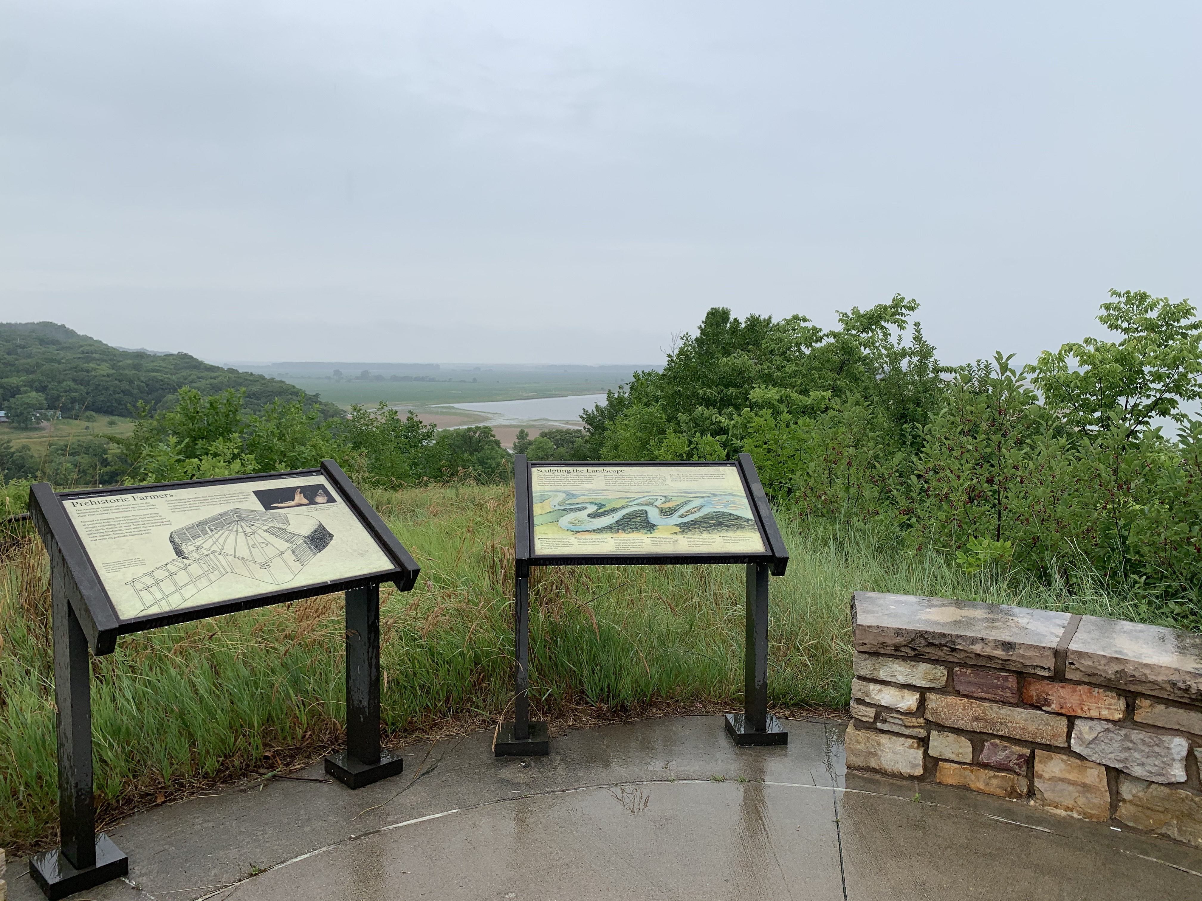 Two waysides and a stone wall overlooking the Missouri River. There are green trees and sandbars in the river.
